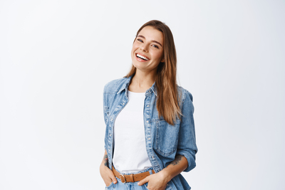 Happy carefree woman with joyful expression, smiling while standing in relaxed pose against white background, holding hands in jeans pockets.